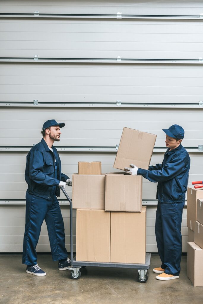 two movers in uniform transporting cardboard boxes on hand truck in warehouse with copy space